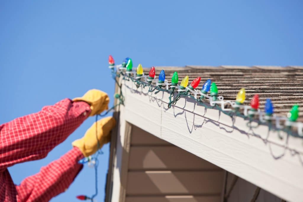 Gloved hands are installing LED Christmas lights onto clips attached to a house roof peak. This image could work equally well for a removing concept. The LEDs are housed within faceted plastic covers which replicate the look of incandescent bulbs. LEDs last longer and are much more efficient than conventional bulbs.