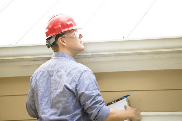 Repairman, building inspector, exterminator, engineer, insurance adjuster, or other blue collar worker has just begun his examination of a building/home's exterior walls, roof, and foundation. He wears a red hard hat and clear safety glasses and holds a his clipboard.